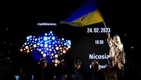 Seorang pengunjuk rasa Ukraina yang tinggal di Siprus, mengibarkan bendera Ukraina ketika yang lain berdiri di depan layar selama protes yang menandai peringatan pertama invasi Rusia ke Ukraina, di ibu kota Nicosia, Siprus. AP/Petros Karadjias