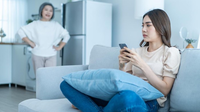 Asian angry senior mature woman shouting at stubborn fussy daughter. Young female sit on sofa use mobile phone chat, disobedient rebellious and ignore annoyed mother. Family problem in house concept.