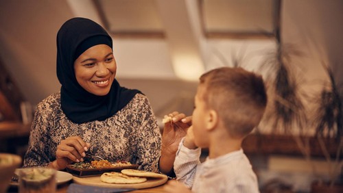 African American Muslim woman enjoying while feeding her son during a meal at dining table.
