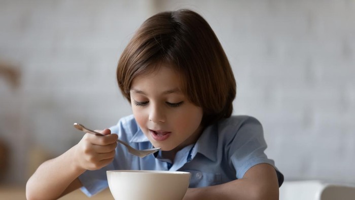 Little hungry cute boy sit at dinner table in kitchen holds spoon eating porridge or whole grain dry cereal with pleasure and appetite, enjoy balanced food. Nutrition, tasty healthy breakfast concept