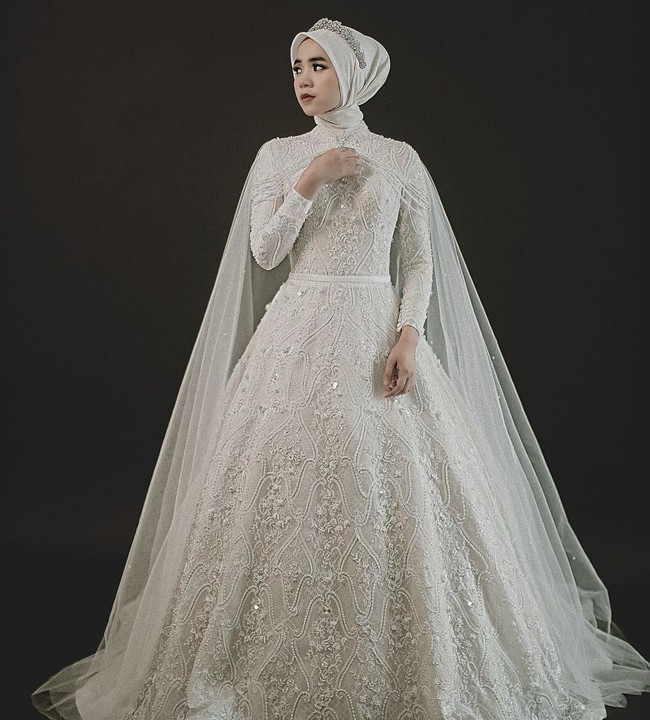 Gaun model ballgown dengan taburan payet, kristal serta bordir membuat tampilan berkesan megah dan cantik. Ditambah cape transparan berbahan sifon yang menambah kesan dramatis. Cocok untuk pernikahan di ballroom atau ruangan berukuran besar. Foto: Weddingku/ Ivory Bridal