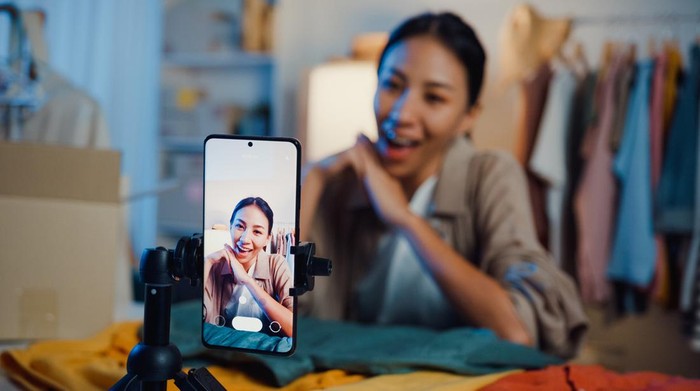 Asian woman live-streamed ecommerce sell clothes in home at night. Beautiful girl using the smartphone and tablet for recording video.
