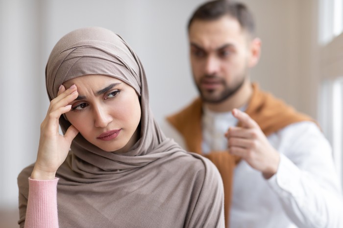 Husband Shouting At Depressed Wife Tired Of Domestic Violence And Victimization Standing At Home. Selective Focus. Relationship With Abuser And Family Conflicts