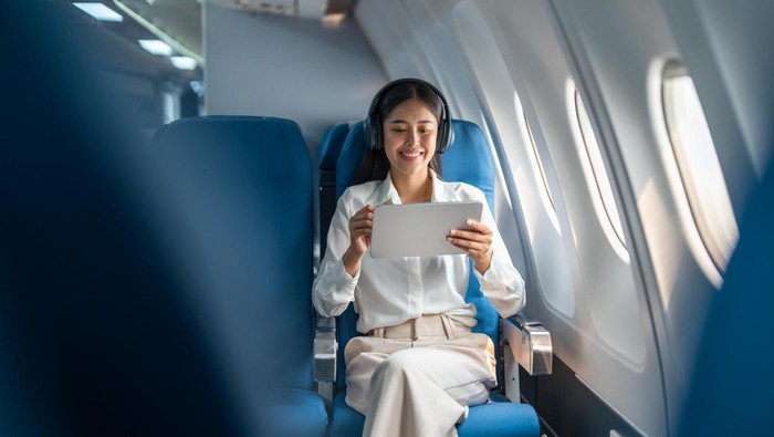 Young Asian business woman or female passenger wearing wireless headphone and working with tablet during the flight.