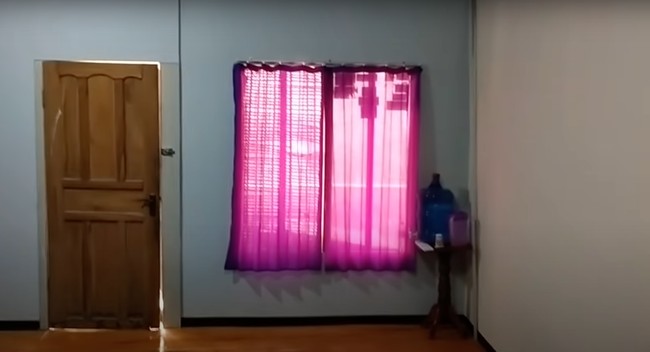 Penampakan bagian dalam rumah menuju ke luar juga terlihat sederhana. Pintu rumah tampak terbuat dari kayu dan gorden jendelanya pun tampak simpel berwarna merah muda. Foto: YouTube/Abiza TV