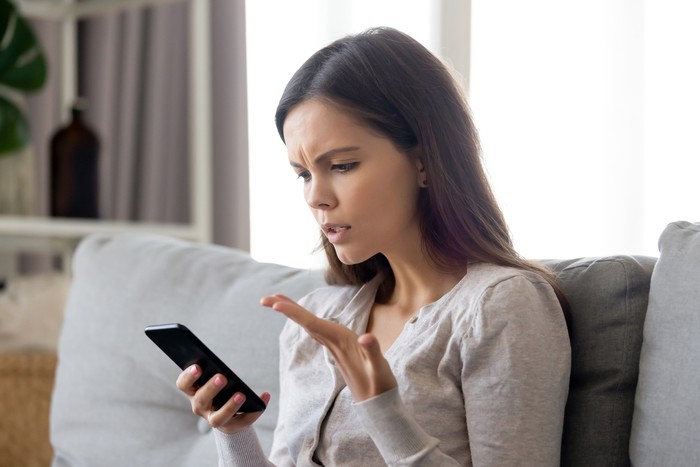 Upset young woman having problem with mobile phone, irritated girl reading bad news in message or email, looking at screen, discharged or broken mobile device, sitting on sofa at home