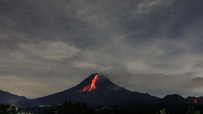 A man rides a motorcycle past a river, while Mount Merapi, a volcanic mountain spews volcanic material in South Kaliurang village, Srumbung district in Central Java, Indonesia on April 30, 2023. Merapi is located in one of the most densely populated parts of Java with over 11,000 people living on the slopes of the mountain. Mount Merapi, measuring 2,968 metres high, is known as one of the most active volcanoes in Indonesia, with an eruption occurring every two to five years. (Photo by Garry Lotulung/NurPhoto via Getty Images)