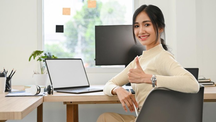 Attractive young Asian businesswoman or female office employee sits at her office desk, showing thump up, smiling and looking at the camera.
