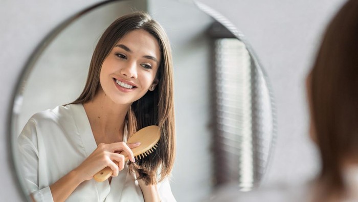 Beauty Routine. Pretty Woman Combing Her Beautiful Hair With Brush While Standing Near Mirror In Bathroom, Attractive Young Lady Looking To Her Reflection And Smiling, Selective Focus With Free Space