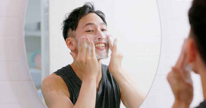 asian man standing in front of mirror is washing his face and he is cheerful