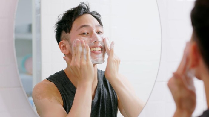 asian man standing in front of mirror is washing his face and he is cheerful