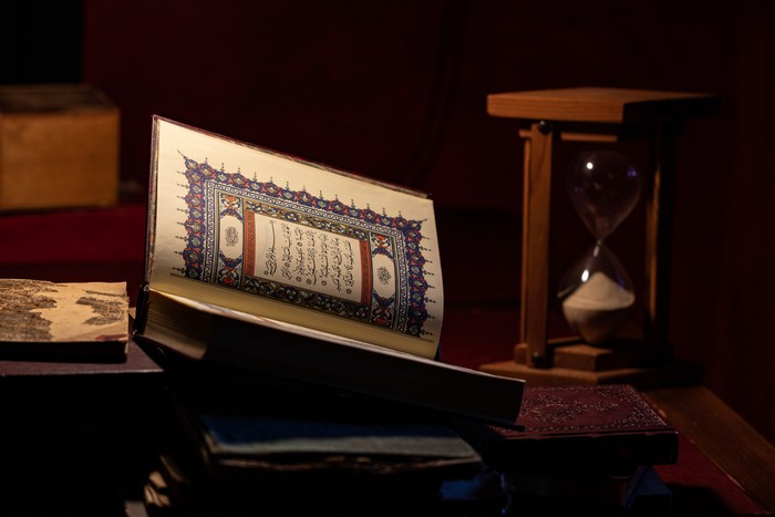Photo of open Holy Book Koran on table by hourglass.  No people are seen in frame. Shot indoor with a full frame mirrorless camera.