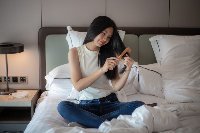 Young beautiful woman is sitting on bed and combing her long brown hair with brush. Home hair care.