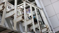 Sekitar tujuh pekerja konstruksi mengenakan rompi warna hijau dan oranye serta helm proyek. Sebagian dari mereka mengelas rangka badan jembatan.  