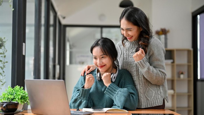 Happy and cheerful Asian businesswomen or business employees clenched fist, looking at laptop screen, celebrating their project success together. victory, winning, good news
