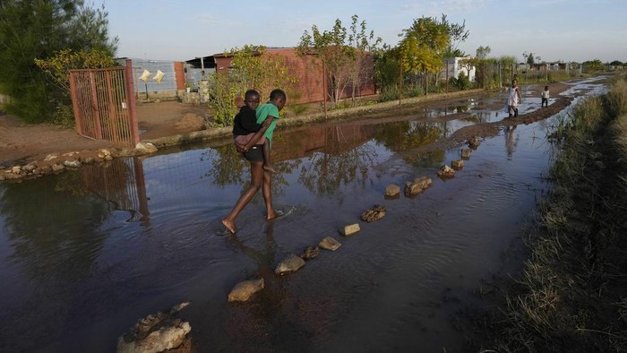 A young girl carries a child on her back through a flooded street by an overflowing reservoir in Hamanskraal, Pretoria, South Africa, Friday, May 26, 2023. Health authorities are yet to confirm the exact source of the cholera outbreak, but poor waste water management and local government instability in South Africa's capital city have been blamed for the situation. (AP Photo/Themba Hadebe)