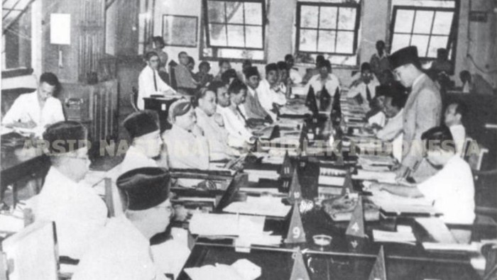 Suasana persidangan PPKI Agustus 1945 untuk menetapkan Undang Undang Dasar 1945, yang pembukaannya menyarikan isi Pancasila, dan memilih presiden dan wakil presiden RI. Tampak Soekarno di kanan tengah (berdiri).