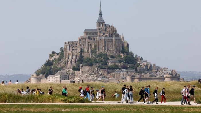 Biara Paling Populer di Prancis Rayakan HUT Ke-1000 People walk past France's Mont-Saint-Michel during the French President's two-day visit in Normandy, in Le Mont-Saint-Michel, north-western France, on June 5, 2023. Emmanuel Macron is on a two-day visit in Normandy as part of preparations to celebrate the millennium of Mont-Saint-Michel Abbey on June 6, for the 80th anniversary of the Allied landings on June 6, 1944, which will take place in 2024. (Photo by Ludovic MARIN / POOL / AFP) (Photo by LUDOVIC MARIN/POOL/AFP via Getty Images)