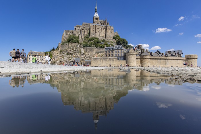 Saint Mont Michel reflecting on water surface during a blue sky day. Mont-Saint-Michel tidal island with the magnificent medieval monastic buildings and the Abbey of Saint Michel with the statue of Archangel Michael atop the spire, in Normandy, France. Le Mont Saint Michel is a UNESCO World Heritage Site since 1979 and second most visited monument in France. The holy island with its breathtaking bay is a major Christian pilgrimage site destination for Europe for centuries. The Mont-Saint-Michel is one of Europes most unforgettable sights. July 2022 (Photo by Nicolas Economou/NurPhoto via Getty Images)