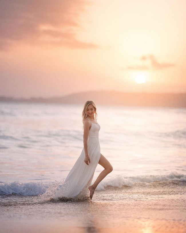 Wanita berambut pirang ini pose bak model dengan latar belakang desiran ombak dan cahaya jingga matahari. Foto: Instagram/@varshaadhikumoro