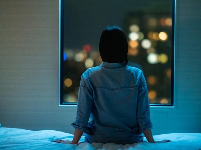 A rear view of a woman sitting alone on a bed in room and looking through the window at night.