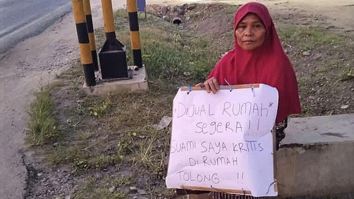 Wanita duduk di pinggir jalan dengan tulisan jual rumah untuk pengobatan suami.