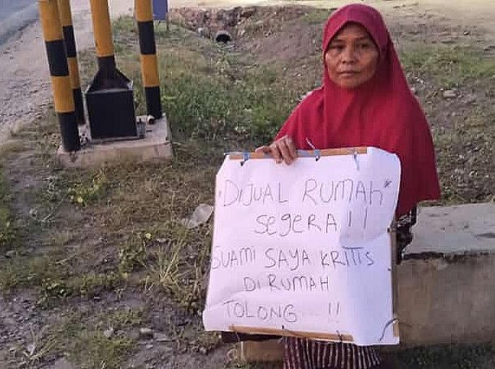 Wanita duduk di pinggir jalan dengan tulisan jual rumah untuk pengobatan suami.