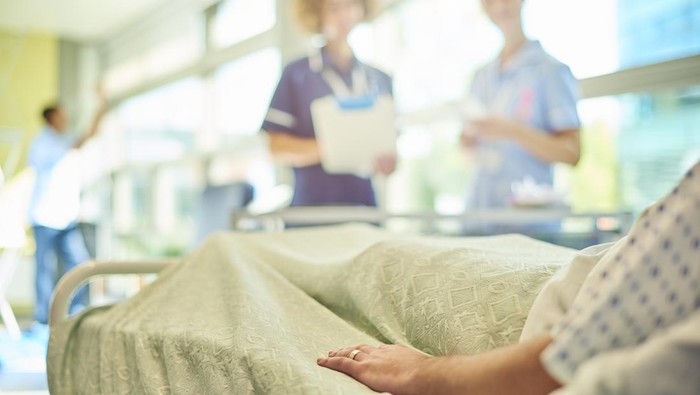 a female senior nurse or matron is  chatting to a young  a young student nurse is attending to the dressings trolley at the foot of a patients bed . They are all defocussed apart from the patients hand in the foreground.