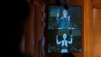 Robot dengan wajah humanoid, pertama-tama membungkuk kepada penonton dan mulai melambaikan tangannya untuk mengontrol tempo pertunjukan langsung.  