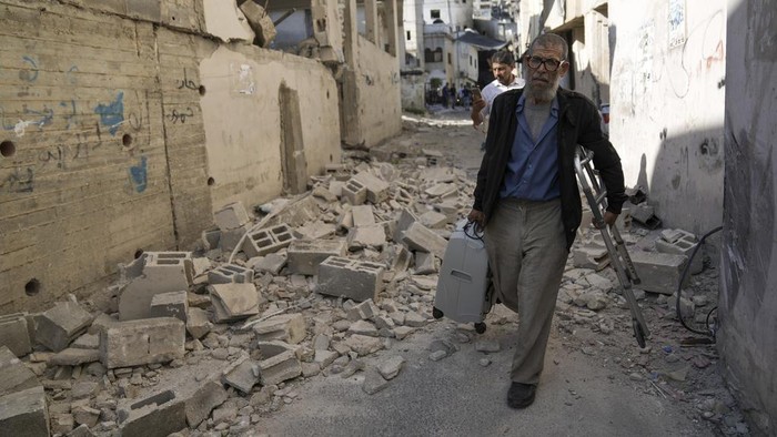 Ramai-ramai Warga Palestina Tinggalkan Jenin yang Dibombardir Israel Palestinians walk by a damaged house in the Jenin refugee camp in the West Bank, Wednesday, July 5, 2023, after the Israeli army withdrew its forces from the militant stronghold. The withdrawal of troops from the camp ended an intense two-day operation that killed at least 13 Palestinians, drove thousands of people from their homes and left a wide swath of damage in its wake. One Israeli soldier was also killed. (AP Photo/Nasser Nasser)