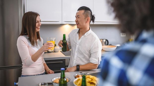 man bun undercut Over the shoulder view of young Asian male and Caucasian female enjoying drinks and conversation in kitchen at social gathering.