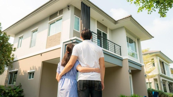 Pasangan berdiri saling merangkul di depan hunian impian mereka yang ingin beli rumah
