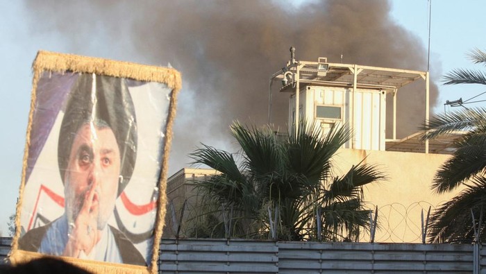 Demonstran Irak Bakar Kedubes Swedia Protes Pembakaran Al-Qur'an A protester holds up a portrait of Iraqi Shi'ite cleric Moqtada al-Sadr as smoke rises from the Swedish embassy building during a protest near the embassy, hours after it was stormed and set on fire ahead of an expected Koran burning in Stockholm, in Baghdad, Iraq, July 20, 2023. (REUTERS/Ahmed Saad)