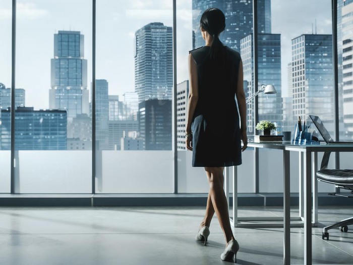 Successful Businesswoman in Stylish Dress Looking out of the Window at Big City in Downtown Area. Confident Female CEO Working on Financial Projects. Manager at Work Planning Marketing Campaign.
