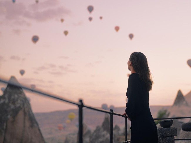 A female tourist is enjoying watching hot air balloons flying in the sky at the rooftop of the hotel where she is staying during her vacation.