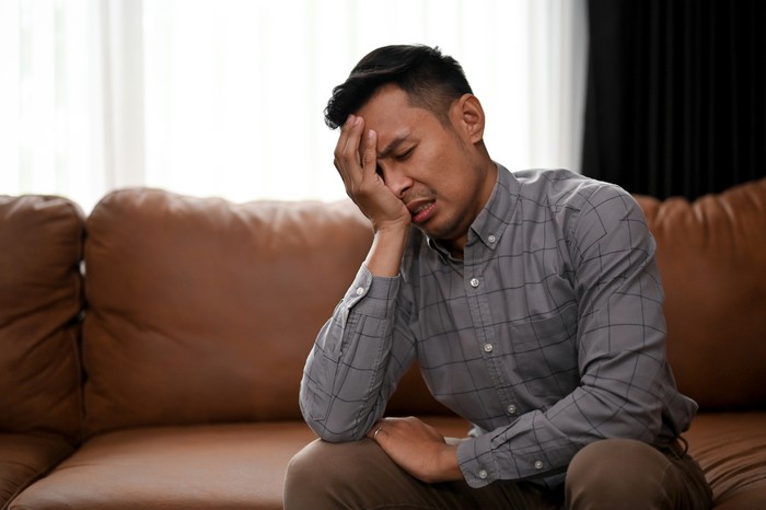 Stressed and upset millennial Asian man is covering his face, crying about his failures in life while sitting in a living room. problem, trouble, broke, depression