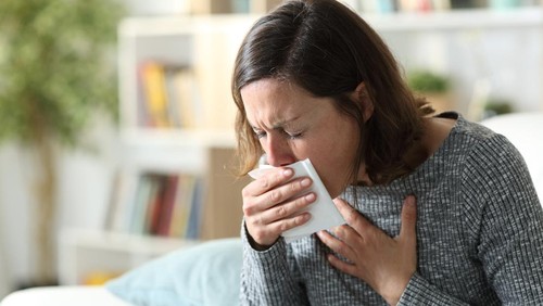 Sick adult woman coughing covering mouth with tissue sitting on a couch at home