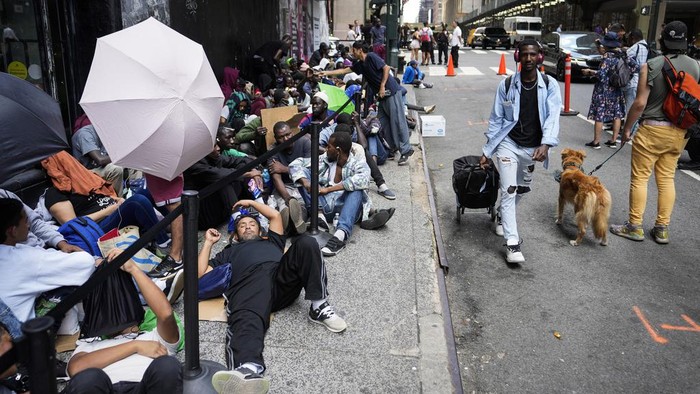 Migrants sit in a queue outside of The Roosevelt Hotel that is being used by the city as temporary housing, Monday, July 31, 2023, in New York. (AP Photo/John Minchillo)