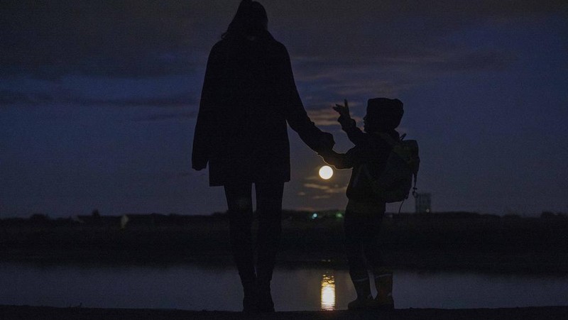 A woman stands with her child in front of a puddle on Tempelhofer Feld and watches the full moon in Berlin Tuesday, Aug. 2, 2023. (Carsten Koall/dpa via AP)