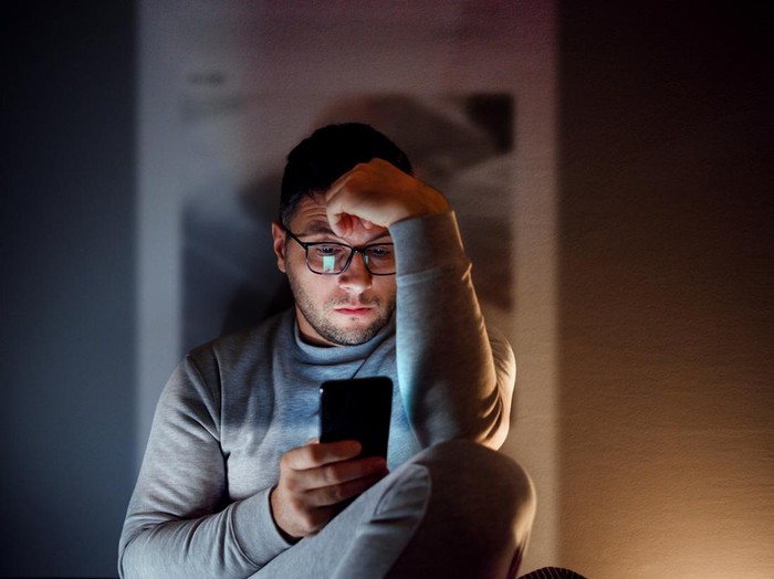 A 35-year-old guy is sitting in the dark room and using his smartphone.