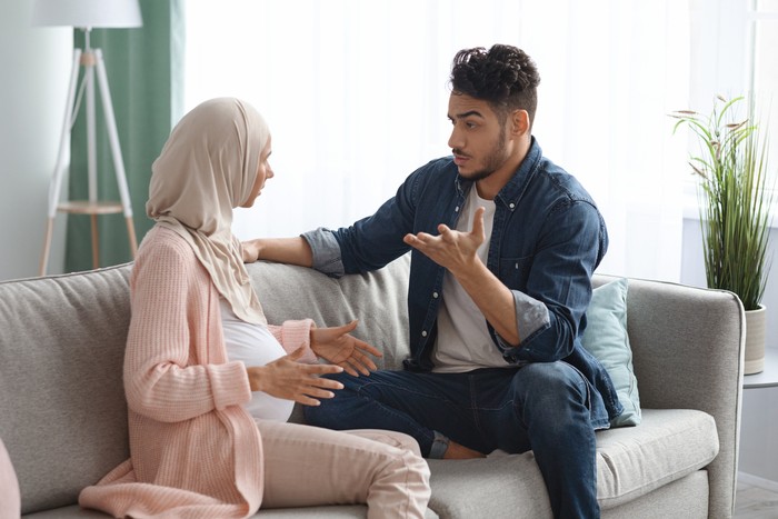 Pregnancy And Relationship Problems. Pregnant Muslim Spouses Arguing At Home, Expectant Muslim Lady In Hijab And Her Arab Husband Quarreling While Sitting On Couch In Living Room, Free Space