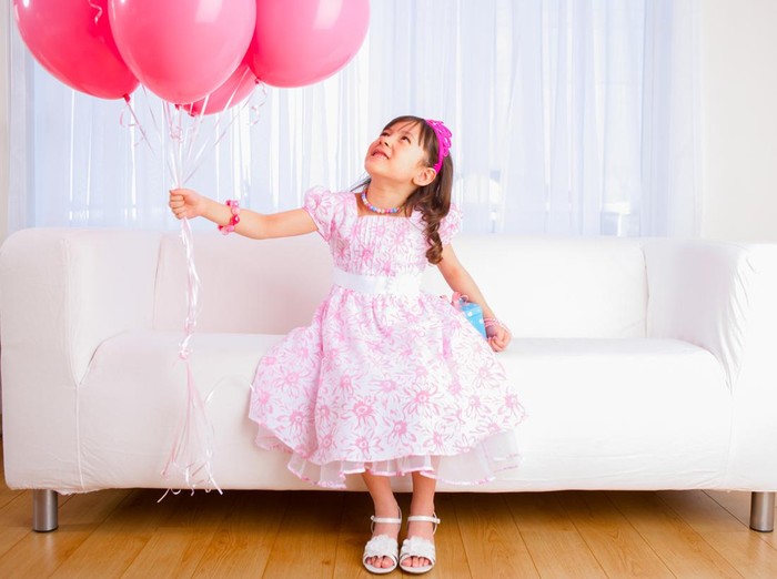 Smiling 8 years girl all dress up celebrating her birthday holding a group of pink balloons sitting on white sofa in livingroom.