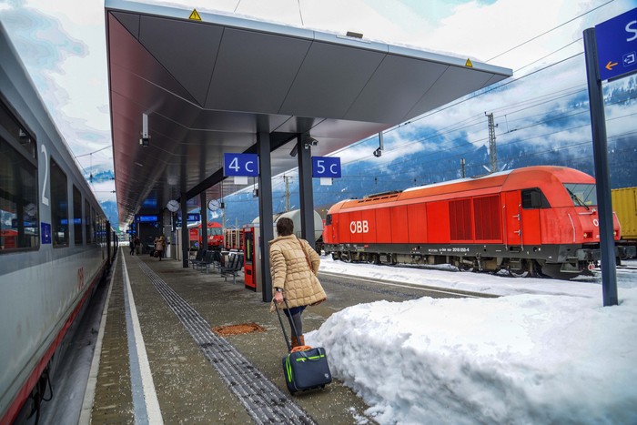 02-21-2022  Salzburg, Austria.  The platform of some station . Passenger with a suitcase on wheelsTrain at left and cargo train - at right- red electric or diesel locomotive