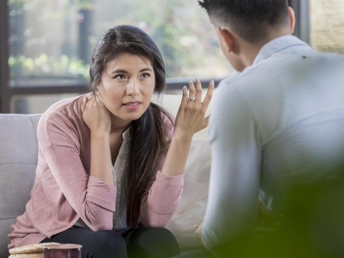 While in a disagreement, a young couple discusses problems. The wife sits on the sofa and gestures in frustration while husband listens intently. The discussion is taking place in a living room.