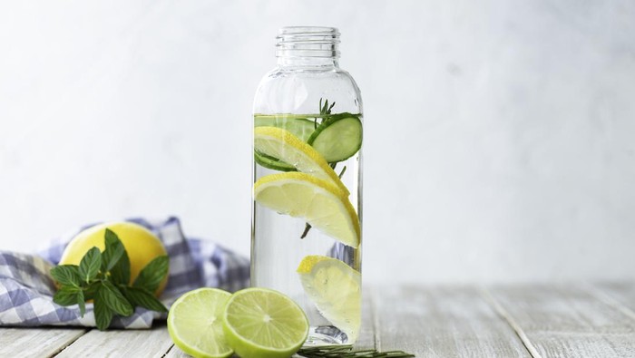 Bottle of infused water on white wood with a slice of lemon , cucumber and rosemary leaf in it.
