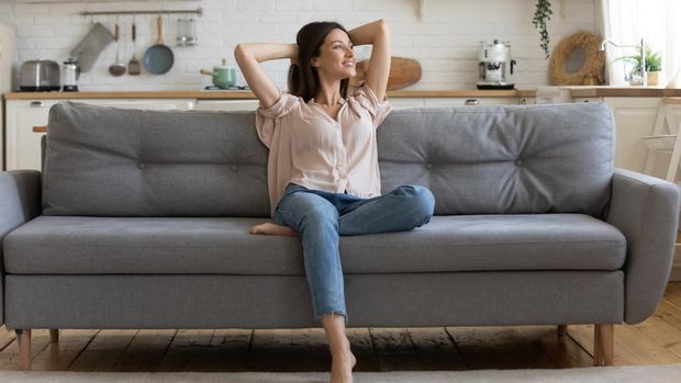 In cozy living room happy woman put hands behind head sitting leaned on couch 30s european female enjoy lazy weekend or vacation, housewife relaxing feels satisfied accomplish chores housework concept