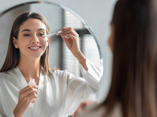 Skin Care Concept. Beautiful Millennial Woman Applying Face Serum Near Mirror, Happy Young Female Standing In Bathroom, Making Daily Skincare Routine At Home, Selective Focus On Reflection