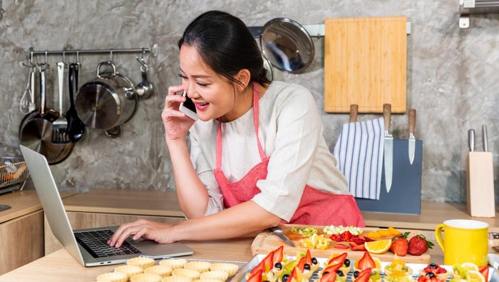 Asian woman making fruit tart for online selling delivery. Female small business bakery shop owner entrepreneur working in laptop computer and mobile phone get order for customer. Small business work at home concept.