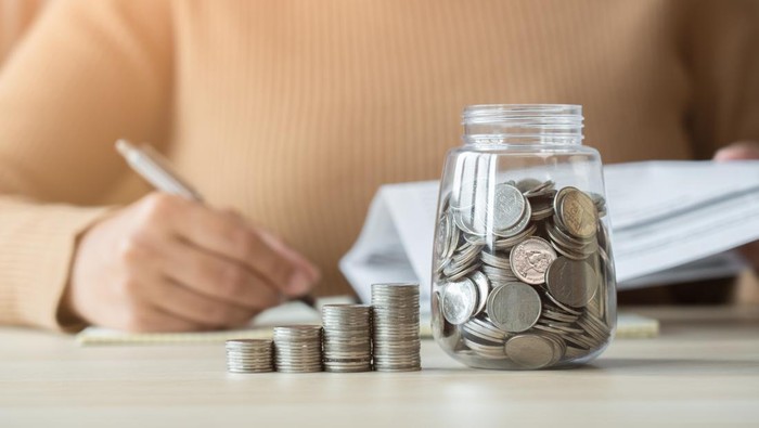 Woman with coins jar taking note to checklist expenses. Concept of doing accounting plan with coin jar and woman writing in a book.