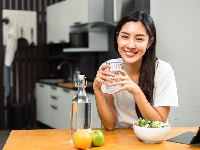 Young female drinking water in glass for refreshing for diet and wellness on table. Beautiful woman happily drink fresh water before breakfast in kitchen in the morning. Healthy food concept.